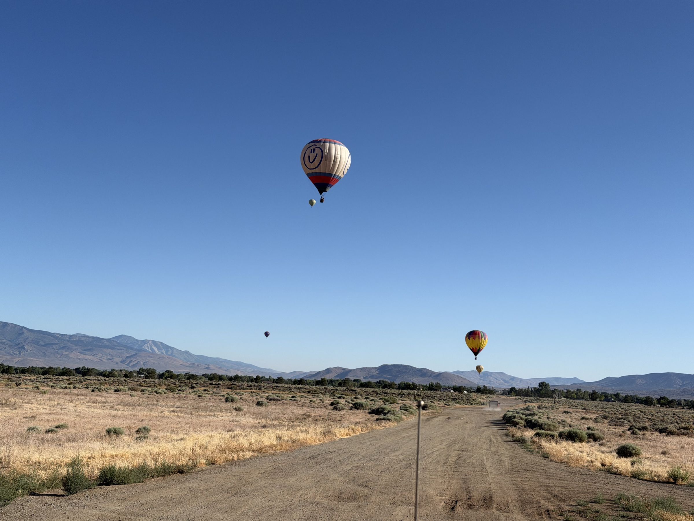 Hot air balloons drifting over open desert scrub and a dirt road at sunrise, with mountains along the horizon