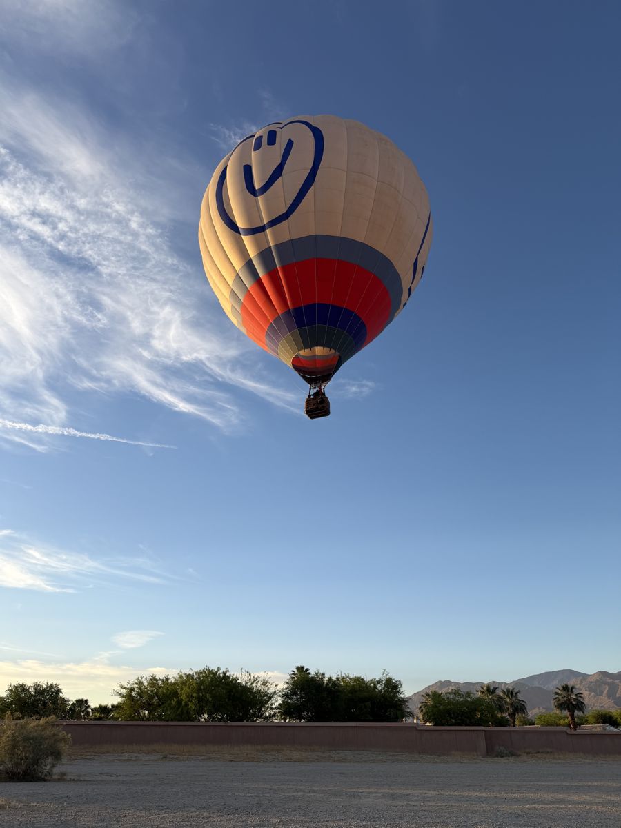 A cream balloon with a smiley face rising above trees and distant hills