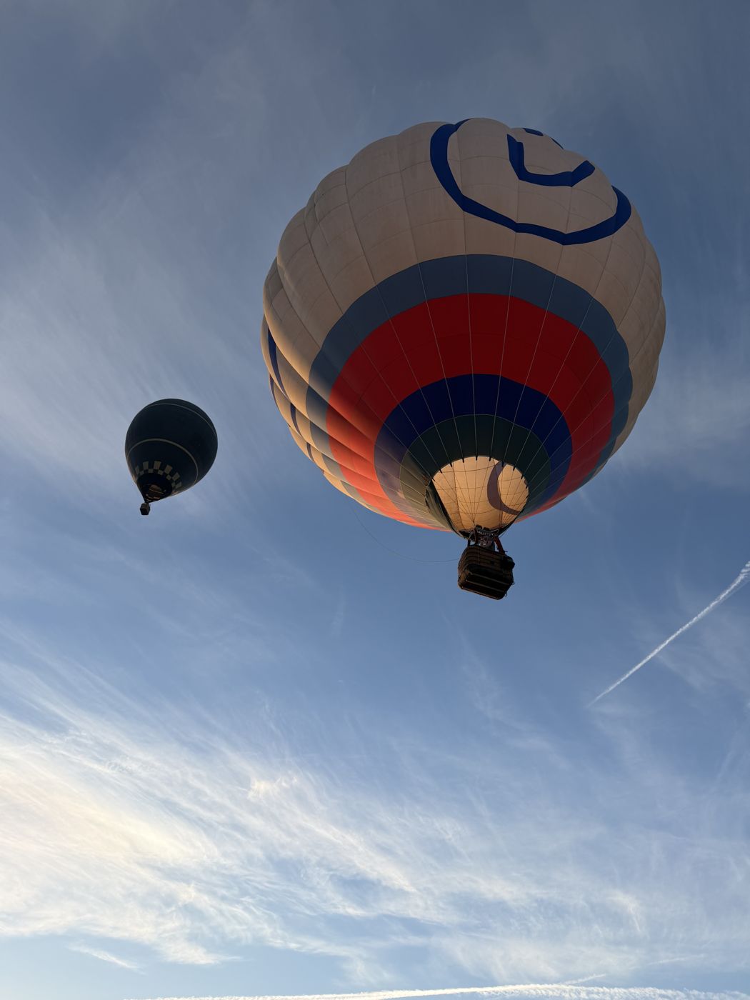 Two hot air balloons climbing into a blue sky streaked with high cloud
